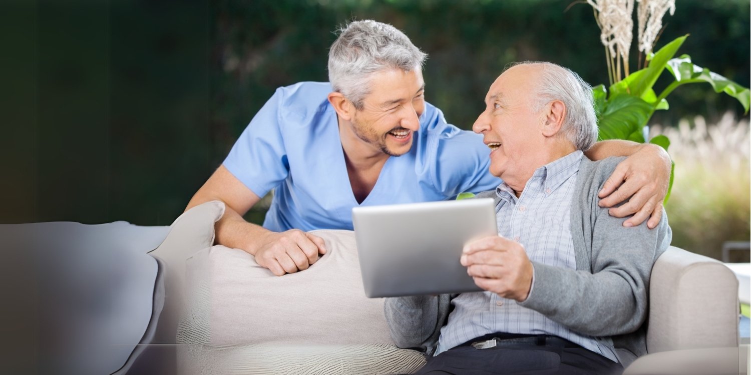 Smiling caregiver with arm around elderly patient holding a tablet.