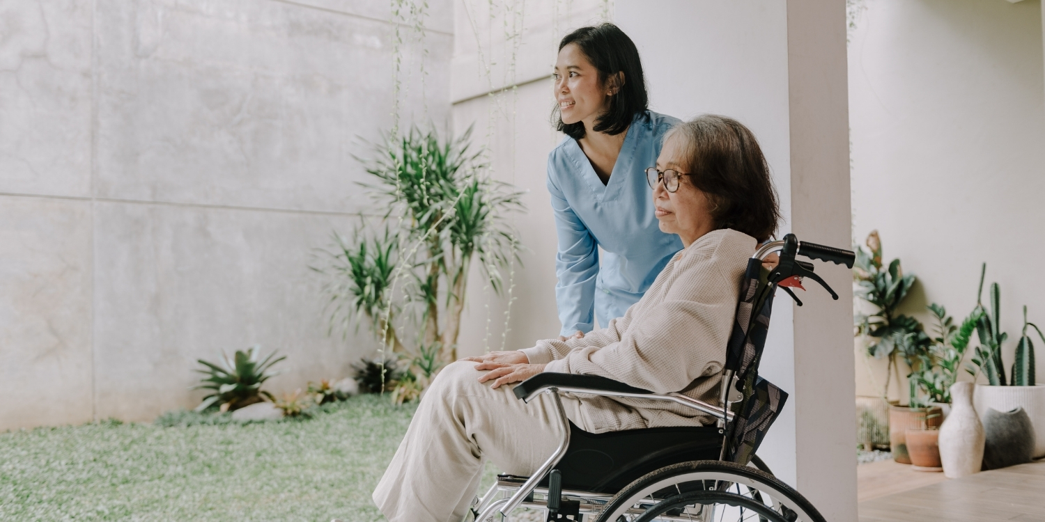 Caregiver standing beside elderly patient in wheelchair.